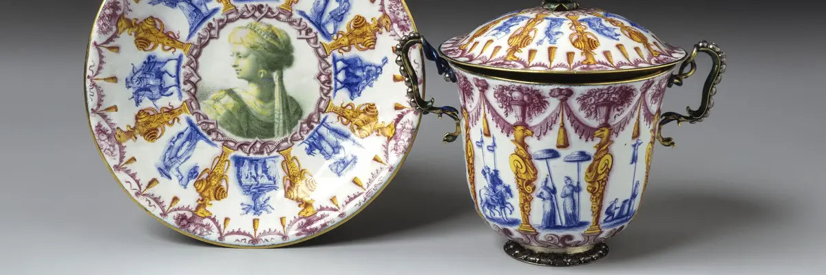 Zu sehen ist eine bunt emaillierte Tasse mit Untertasse mit chinesisch anmutendem Dekor. Hierzu gehören zum Beispiel zwei Figuren mit großen Sonnenschirmen, zwei Fabelwesen sowie Girlanden und Ornamente.