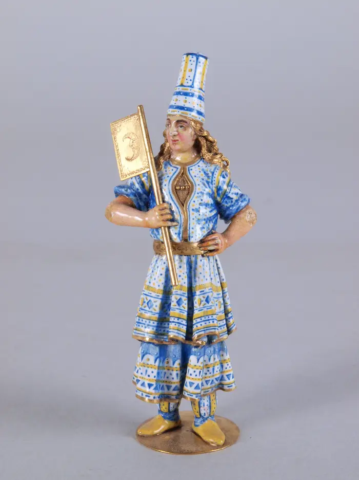 Diese Abbildung zeigt eine kleine Figur. Sie trägt eine blau verzierte Fantasie-Tracht und hält eine Fahne in der Hand.