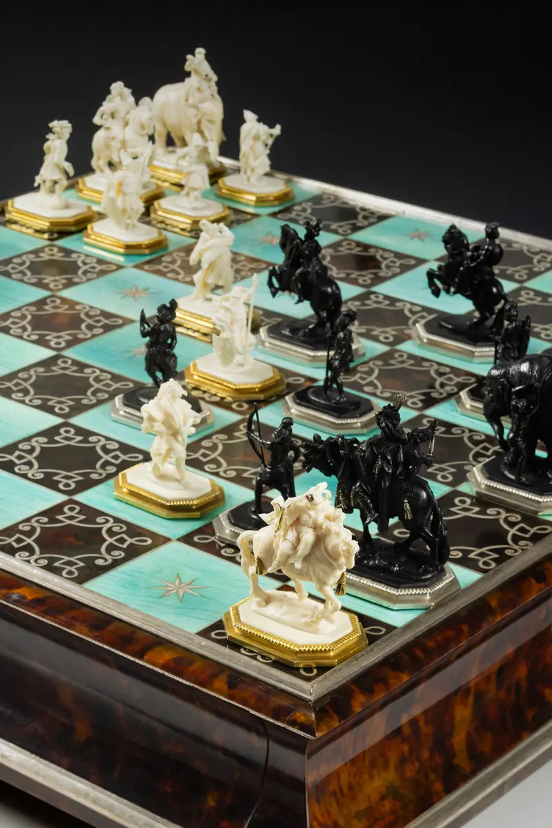 Hier ist ein kunstvoll gearbeitetes Schachspiel zu sehen. Das Schachbrett ist in türkise und braune Spielfelder unterteilt. Die Figuren sind kunstvoll aus hellem Elfenbein und dunklem Ebenholz geschnitzt.