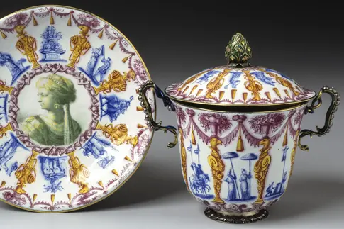 Zu sehen ist eine bunt emaillierte Tasse mit Untertasse mit chinesisch anmutendem Dekor. Hierzu gehören zum Beispiel zwei Figuren mit großen Sonnenschirmen, zwei Fabelwesen sowie Girlanden und Ornamente.