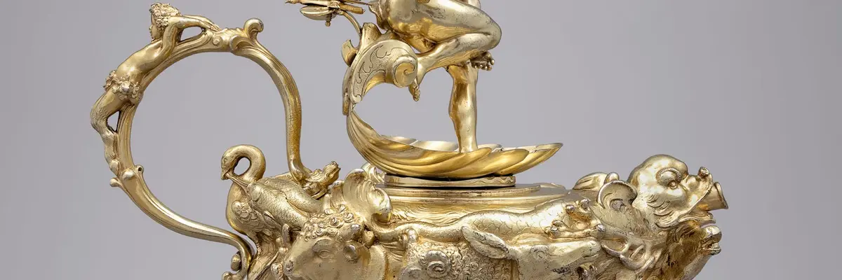 Die Abbildung zeigt eine reich verzierte goldene Kanne mit einem sehr dünnen Henkel. Auf dem Deckel befindet sich eine Figur.