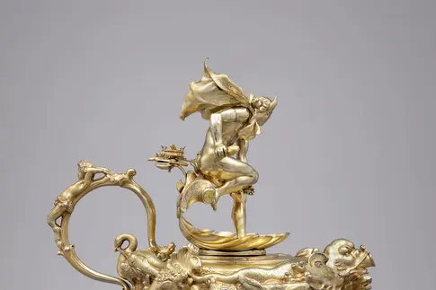 Die Abbildung zeigt eine reich verzierte goldene Kanne mit einem sehr dünnen Henkel. Auf dem Deckel befindet sich eine Figur.